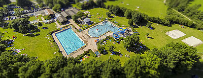 Luftaufnahme eines Freibads mit Schwimmbecken, Planschbecken mit Wasserrutsche und Liegewiese mit Sonnenschirmen und Menschen Luftaufnahme eines Freibads mit Schwimmbecken, Planschbecken mit Wasserrutsche und Liegewiese mit Sonnenschirmen und Menschen