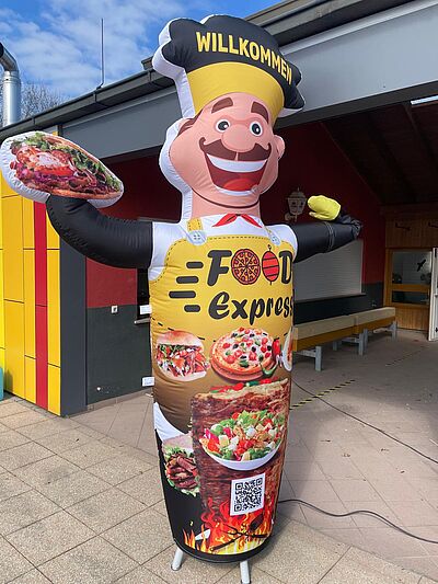 Aufblasbare Werbefigur mit Kochmütze und Schrift 'Willkommen', hält Pizza und wirbt für 'Food Express' vor einem Gebäude